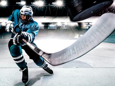 Hockey player in full gear takes a powerful swing with his stick towards a black puck floating in the air. Banks of arena lights illuminate the night sky.