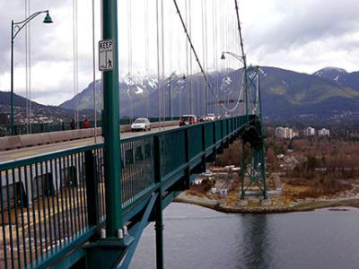 Against a background of mountains, cars cross a suspension bridge that takes them to the city of Vancouver BC.